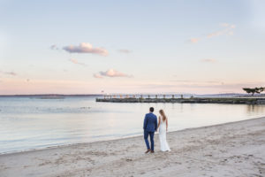 A bride and groom walk on the beach at sunset on their wedding day at Shenorock Shore Club in Rye, New York. Photo by Kelly Kollar Photography.